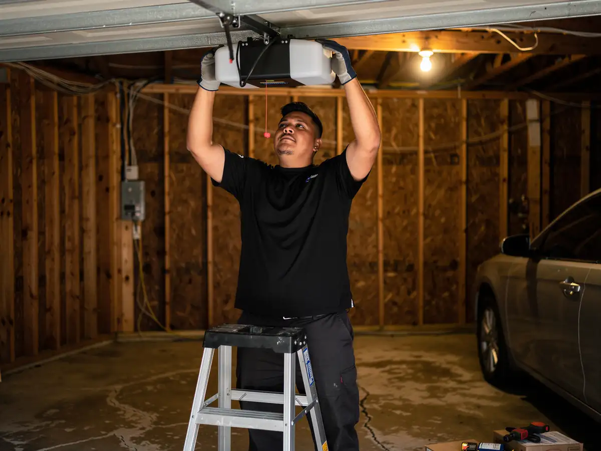 A technician on a ladder installs a garage door opener in a garage.